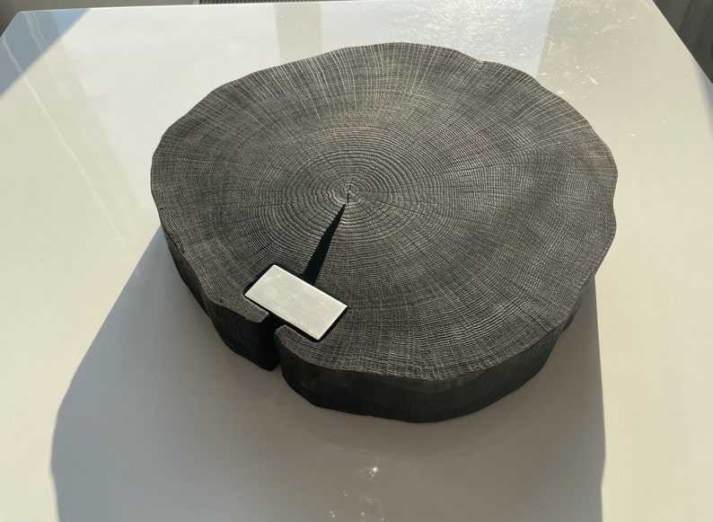 May include: A large, round, black wooden serving board with a natural wood grain pattern. The board has a small, rectangular, silver metal insert in the center.