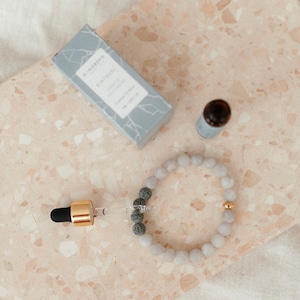 Könnte beinhalten: Ein graues Lava-Stein-Armband mit einem goldenen Perlenakzent. Das Armband liegt auf einer hellbraunen Oberfläche mit einer kleinen Flasche ätherischem Öl und einer Schachtel mit der Aufschrift "By NORDEN" und dem Text "KIN-TSUGI BALANCE RECOVERY" auf der Vorderseite.