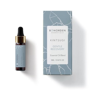 Könnte beinhalten: Eine kleine, durchsichtige Glasflasche mit goldenem Deckel und einem Etikett mit der Aufschrift "BONORDEN ESSENTIALS KINTSUGI GENTLE RECOVERY Essential Oil Blend 5ML / 0.16 FL OZ". Die Flasche steht auf einer weißen Oberfläche. Der Hintergrund ist hellblau mit einem weißen Muster.