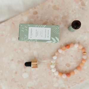 Könnte beinhalten: Draufsicht auf eine hellgrüne Schachtel und eine kleine Glasflasche, beide mit minimalistischem Design. Auf der Schachtel steht "SHINRIN YOKU Inner Balance Essential Oil Blend". Ein Armband mit Pfirsich- und weißen Perlen ist ebenfalls vorhanden.