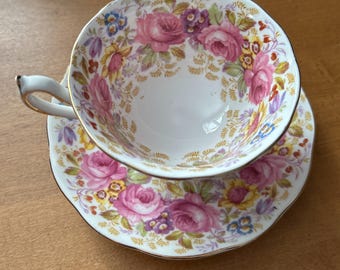 Puede incluir: Un juego de taza y platillo de té vintage con un patrón floral. La taza y el platillo son blancos con un borde dorado y presentan flores rosas, amarillas y azules.