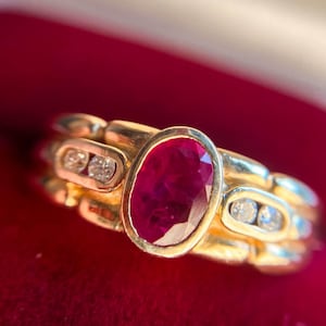 Puede incluir: Un anillo de oro con una gran piedra de rubí ovalada rodeada de diamantes más pequeños. El anillo está engastado en una banda de oro con un diseño texturizado.