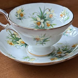May include: A white teacup and saucer with a floral pattern of yellow daffodils and green leaves. The cup and saucer have a gold trim.