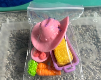 May include: A pink cowboy hat, a purple dish with a yellow food item, a green and white dish with red and orange food items, and a green and yellow vegetable in a clear plastic bag.