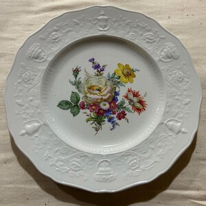 May include: White porcelain plate with a scalloped edge and embossed floral and scrollwork details. The center features a colorful floral bouquet with yellow, pink, and purple flowers. A decorative piece for serving or display.