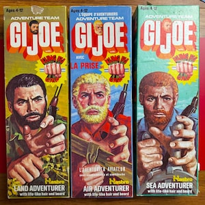 Pode incluir: Três caixas de brinquedos retangulares G.I. Joe Adventure Team com ilustrações de aventureiros terrestres, aéreos e marítimos com cabelo e barba realistas. Cada caixa exibe o logotipo G.I. Joe, o texto Kung Fu Grip e a idade 4-12.