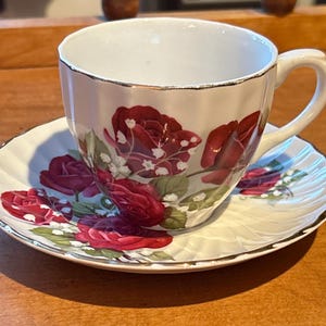 Peut inclure: Une tasse à thé et une soucoupe blanches avec une bordure dorée et un motif floral de roses rouges et de fleurs blanches.