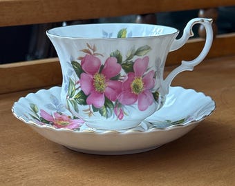 Puede incluir: Un juego de taza y platillo de té blanco con un diseño floral. La taza tiene un asa delicada y un borde festoneado. El platillo tiene un borde festoneado a juego y un diseño floral. Las flores son rosas y blancas con hojas verdes.