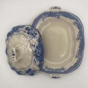 Può includere: Un piatto da portata in ceramica color crema con coperchio abbinato, caratterizzato da un motivo floreale blu e un bordo a righe. Il piatto rettangolare ha due manici laterali e un pomello centrale sul coperchio, decorato con motivi botanici blu.