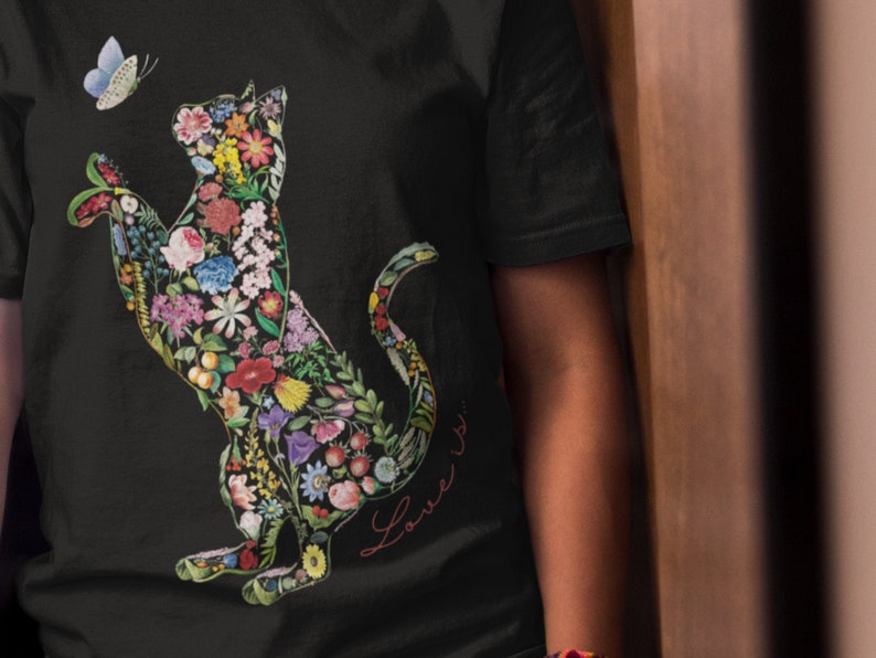 May include: Black t-shirt featuring a floral cat design with a butterfly. The cat silhouette is filled with colorful flowers and greenery. The words "Love is..." are written in cursive on the shirt.