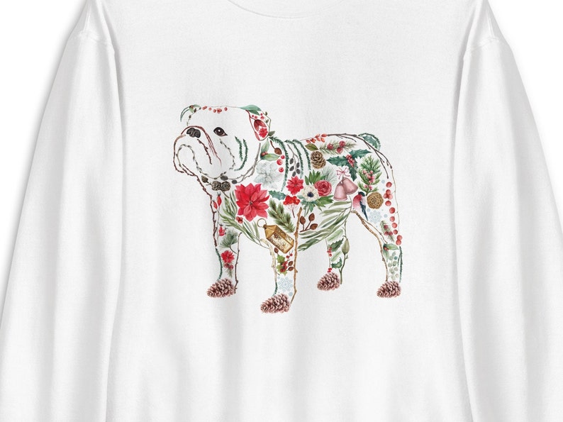 Peut inclure: Sweat-shirt blanc avec une illustration &agrave; l'aquarelle d'un bouledogue orn&eacute; de feuillage de No&euml;l. Le bouledogue est orn&eacute; de poinsettias rouges, de feuilles vertes et de pommes de pin, cr&eacute;ant un design de vacances festif.
