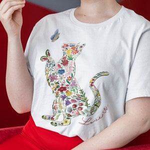 May include: White t-shirt featuring a cat silhouette filled with colorful flowers and foliage. A blue butterfly hovers near the cat's head. The words "Love is..." are written in red script below the cat design. The shirt is made of a soft material.