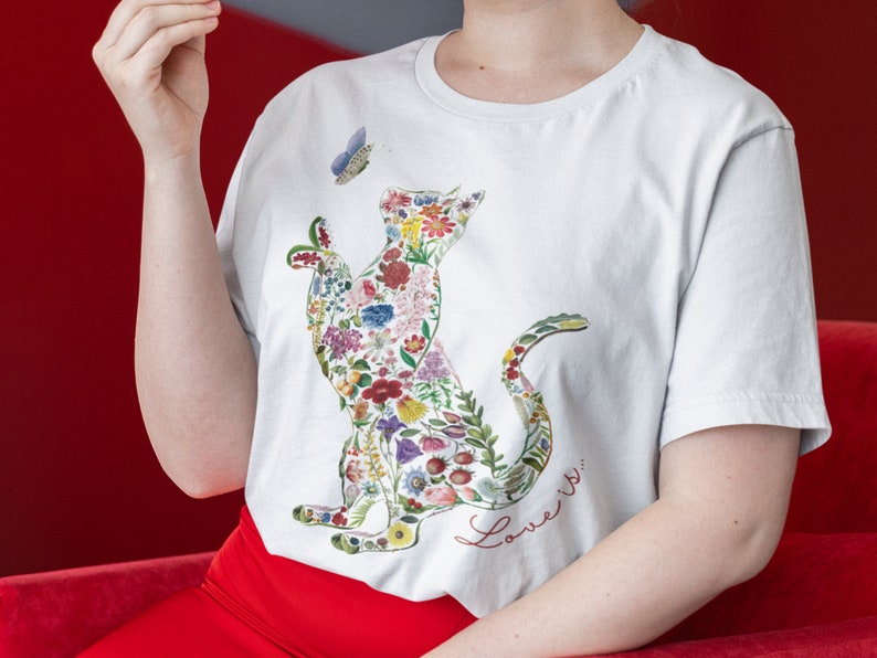 May include: White t-shirt featuring a cat silhouette filled with colorful flowers and foliage. A blue butterfly hovers near the cat's head. The words "Love is..." are written in red script below the cat design. The shirt is made of a soft material.