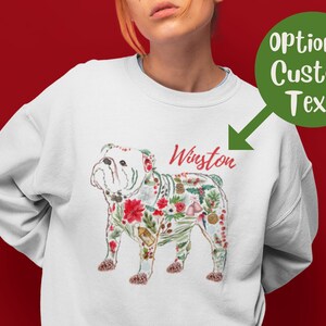 Peut inclure: Sweat-shirt blanc avec un bouledogue floral et le nom "Winston" en script rouge. Le bouledogue est orn&eacute; de poinsettias rouges, de feuilles vertes et d'&eacute;l&eacute;ments festifs. Un cercle vert avec "Optional Custom Text" est en haut &agrave; droite.