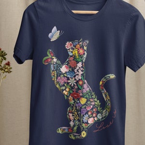 May include: Navy blue t-shirt featuring a floral cat design with a butterfly. The cat is filled with colorful flowers and foliage. The words "Love is..." are written in cursive script. The shirt is hanging on a wooden hanger.