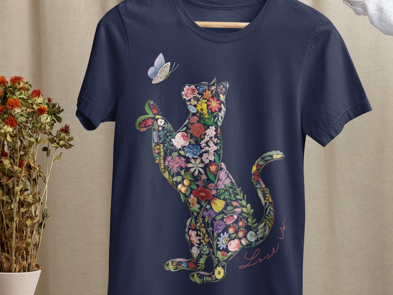 May include: Navy blue t-shirt featuring a floral cat design with a butterfly. The cat is filled with colorful flowers and foliage. The words "Love is..." are written in cursive script. The shirt is hanging on a wooden hanger.