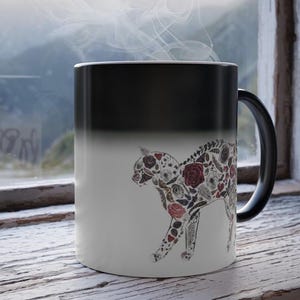 Puede incluir: Una taza que cambia de color en blanco y negro con un diseño de gato lleno de elementos florales y botánicos en rojo, blanco y marrón. La taza está colocada frente a una ventana con una vista exterior borrosa.