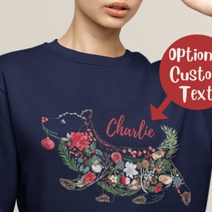 Może przedstawiać: Bordowy sweter z kwiatowym wzorem w kształcie psa. Wzór zawiera czerwone poinsecje, zielone liście i małego ptaka. Tekst "Charlie" jest nadrukowany nad psem.