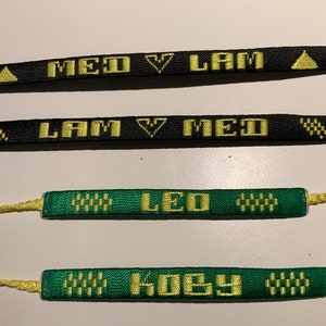 Op de afbeelding: Vier geweven vriendschapsarmbanden met gele letters op zwarte en groene banden. De armbanden lezen "MED V LAM", "LAM V MED", "LEO" en "KOBEY".