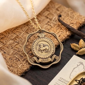 May include: A gold-colored pendant necklace featuring a circular medallion with a lion emblem, a crown, and text. The pendant hangs from a delicate chain. The necklace is displayed on a textured surface with other items.