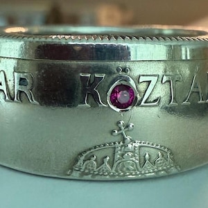 May include: A silver ring with the words "MAGYAR KÖZTARSASAG" engraved around the band. A small, round, pink gemstone is set into the ring. A crown design is visible below the gemstone.
