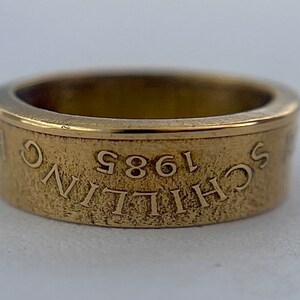 K&ouml;nnte beinhalten: Ein goldfarbener Metallring mit strukturierter Oberfl&auml;che. Das &auml;u&szlig;ere Band zeigt den eingravierten Text "SCHILLING 1985", der auf dem Kopf steht. Der Ring hat einen glatten, polierten Innenrand und ein flaches, breites Bandprofil mit metallischem Glanz.