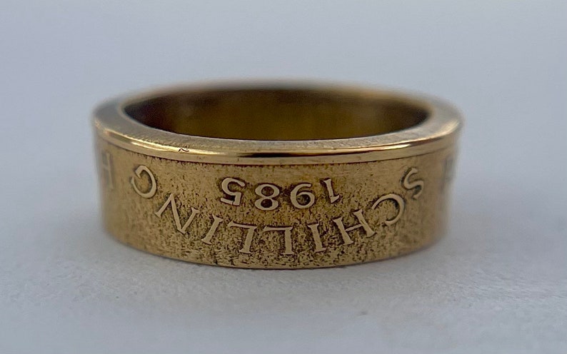 K&ouml;nnte beinhalten: Ein goldfarbener Metallring mit strukturierter Oberfl&auml;che. Das &auml;u&szlig;ere Band zeigt den eingravierten Text "SCHILLING 1985", der auf dem Kopf steht. Der Ring hat einen glatten, polierten Innenrand und ein flaches, breites Bandprofil mit metallischem Glanz.