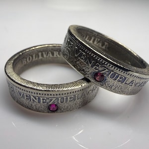 May include: Two silver-toned coin rings crafted from Venezuelan currency, each featuring the embossed text "BOLIVAR" and "VENEZUELA" along the outer band. Each ring is accented with a single, small, circular, deep pink gemstone set into the metal.