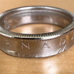 Może przedstawiać: Srebrny pierścionek wykonany z kanadyjskiej monety. Na boku pierścionka wytłoczony jest napis "CANADA". Pierścionek ma teksturowaną krawędź i gładkie wnętrze. Pierścionek leży na jasnobrązowej powierzchni.
