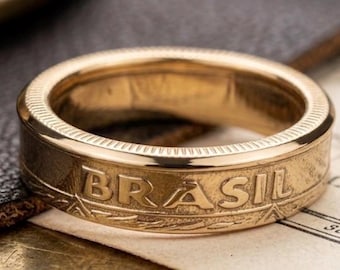 May include: A gold-toned metal ring featuring a band with a reeded edge and the word BRASIL embossed in capital letters on the outer surface. The ring has a polished finish and a flat profile, showcasing a coin-like design with intricate detailing.