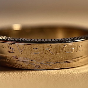 May include: A silver ring with the word "SVERIGE" engraved on the band. The ring has a textured edge and a detailed design. The ring is a close-up shot, highlighting the craftsmanship and the inscription.