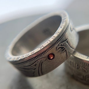 Può includere: Due anelli d'argento con un designo dettagliato e una pietra rossa su ciascuno. Gli anelli sono realizzati con monete riutilizzate.
