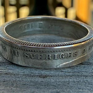 May include: A silver ring with engraved text, possibly a repurposed coin. The ring has a textured edge and visible markings. The ring is displayed on a weathered wooden surface.