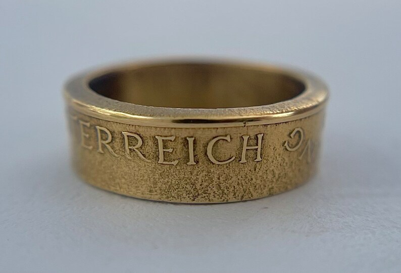 K&ouml;nnte beinhalten: Ein goldfarbener Metallring mit einem glatten, polierten oberen Rand und einem strukturierten Au&szlig;enband. Die Au&szlig;enfl&auml;che zeigt den gepr&auml;gten Text "ERREICH", der in einer Serifenschrift deutlich sichtbar um den Umfang des Rings verl&auml;uft.