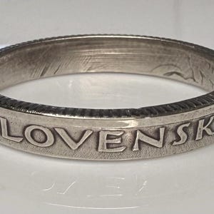 Puede incluir: Un anillo de plata con la palabra "SLOVENSKA" grabada alrededor de la banda. El anillo tiene un borde texturizado y una apariencia ligeramente desgastada, lo que sugiere que puede ser vintage o hecho a mano. El anillo se muestra sobre una superficie blanca.