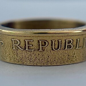 K&ouml;nnte beinhalten: Ein goldfarbener Metallring mit strukturierter Bandoberfl&auml;che. Die Au&szlig;enseite des Rings zeigt die gepr&auml;gten Gro&szlig;buchstaben REPUBLIK, mit einem glatten, polierten oberen Rand und einem etwas dunkleren, mattierten Bandk&ouml;rper.