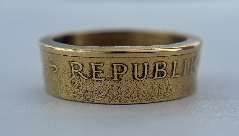 K&ouml;nnte beinhalten: Ein goldfarbener Metallring mit strukturierter Bandoberfl&auml;che. Die Au&szlig;enseite des Rings zeigt die gepr&auml;gten Gro&szlig;buchstaben REPUBLIK, mit einem glatten, polierten oberen Rand und einem etwas dunkleren, mattierten Bandk&ouml;rper.
