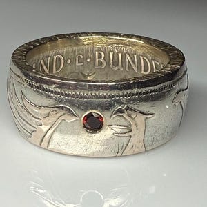 May include: Silver ring with engraved bird designs and a small, round, red gemstone. The ring has a wide band with the inscription "BUNDE" visible. A unique piece of jewelry.
