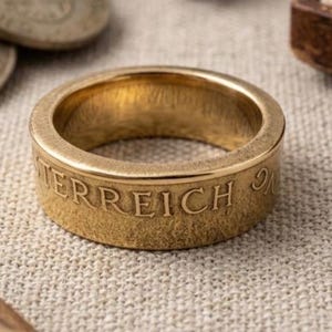 K&ouml;nnte beinhalten: Ein goldfarbener Metallring, der aus einer M&uuml;nze gefertigt wurde, mit einer glatten, polierten Innenseite und einem strukturierten Au&szlig;enband. Das Wort OSTERREICH ist in Gro&szlig;buchstaben auf der Au&szlig;enfl&auml;che des Rings eingepr&auml;gt.