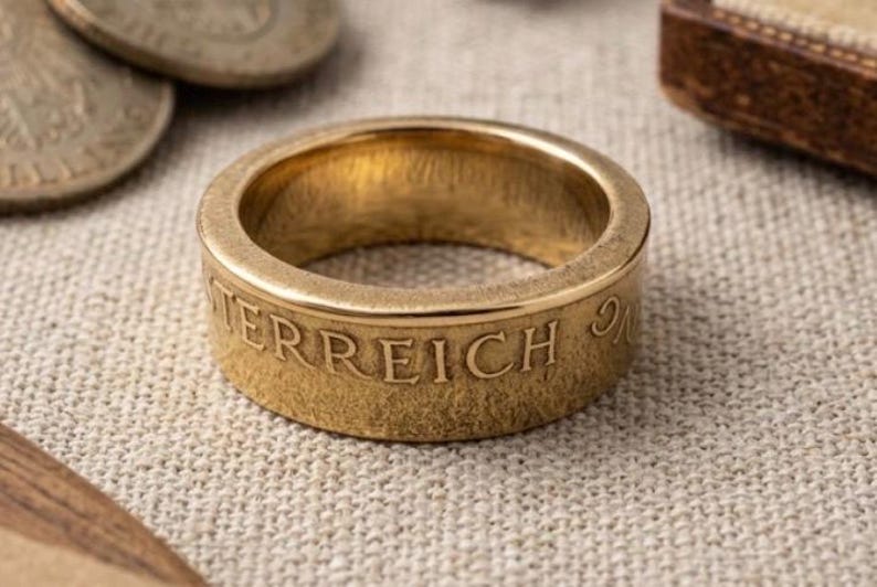 K&ouml;nnte beinhalten: Ein goldfarbener Metallring, der aus einer M&uuml;nze gefertigt wurde, mit einer glatten, polierten Innenseite und einem strukturierten Au&szlig;enband. Das Wort OSTERREICH ist in Gro&szlig;buchstaben auf der Au&szlig;enfl&auml;che des Rings eingepr&auml;gt.