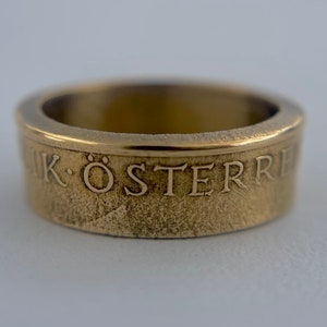 K&ouml;nnte beinhalten: Ein goldfarbener Metallring mit einer texturierten Oberfl&auml;che und einem glatten oberen Rand. Die Au&szlig;enseite des Bandes zeigt den gepr&auml;gten Text "IK &Ouml;STERRE" in einer Serifenschrift, der sich um den Umfang des kreisf&ouml;rmigen Schmuckst&uuml;cks zieht.