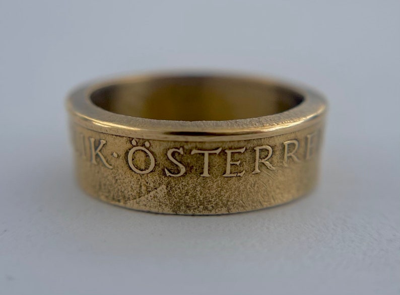 K&ouml;nnte beinhalten: Ein goldfarbener Metallring mit einer texturierten Oberfl&auml;che und einem glatten oberen Rand. Die Au&szlig;enseite des Bandes zeigt den gepr&auml;gten Text "IK &Ouml;STERRE" in einer Serifenschrift, der sich um den Umfang des kreisf&ouml;rmigen Schmuckst&uuml;cks zieht.