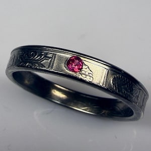 May include: A silver ring with a dark, textured finish. The ring has an engraved pattern along the band and a small, round, bright pink gemstone set in the centre. The ring is a piece of jewellery.