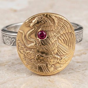 May include: A gold-colored ring with a detailed emblem featuring an eagle, snake, and cactus design, with a small red gemstone. The ring has a silver band with an engraved pattern. Jewelry.