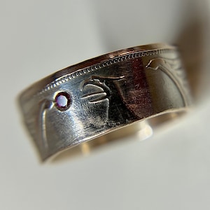 May include: Silver ring with a red gemstone set into the band. The ring is made from a repurposed coin with a design on the band.