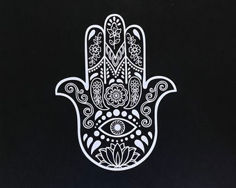 May include: A white hamsa hand with intricate floral designs and an eye in the center, against a black background.
