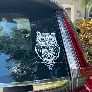 May include: White owl decal with blue eyes on a black car window. The owl is stylized with intricate details and a tribal design. The decal has the text "Amaryllis Expressions" at the bottom.