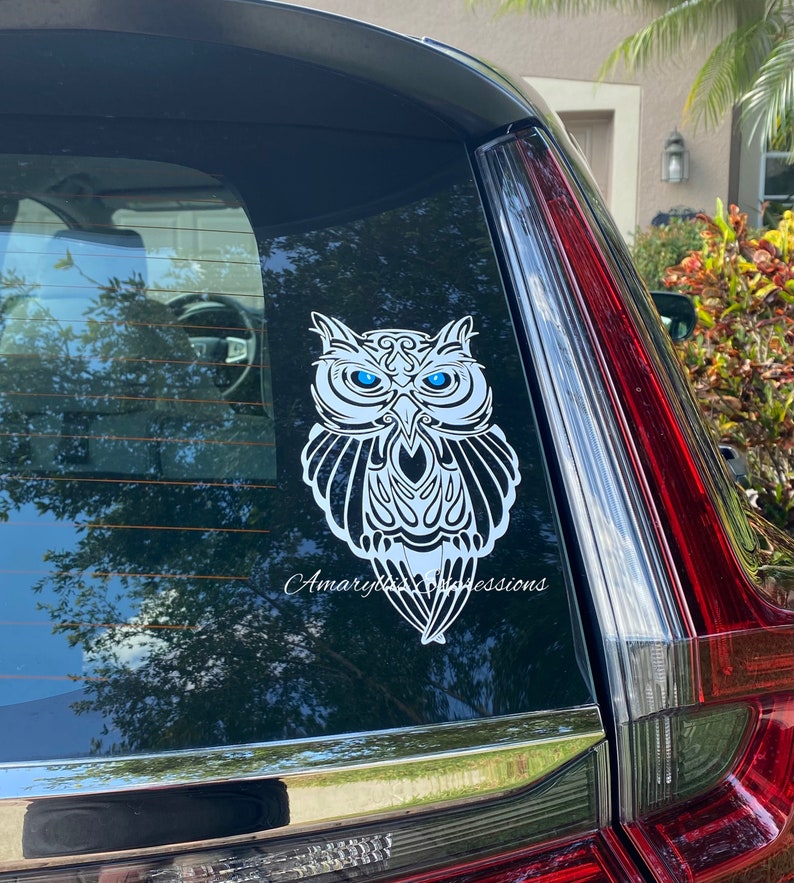 May include: White owl decal with blue eyes on a black car window. The owl is stylized with intricate details and a tribal design. The decal has the text "Amaryllis Expressions" at the bottom.