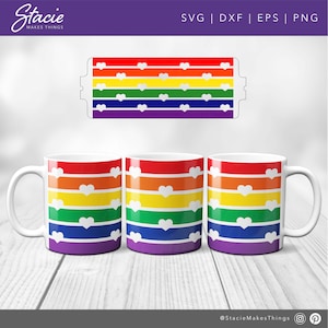 May include: Three white mugs with a rainbow stripe design and white hearts. The stripes are red, orange, yellow, green, blue, indigo, and violet. The mugs are on a white wood surface.