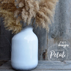 May include: A white ceramic pitcher with a black ombre effect at the bottom, filled with dried pampas grass. The pitcher is sitting on a wooden surface. The text "TWO designs SUGAR Petal designs" is visible in the background.